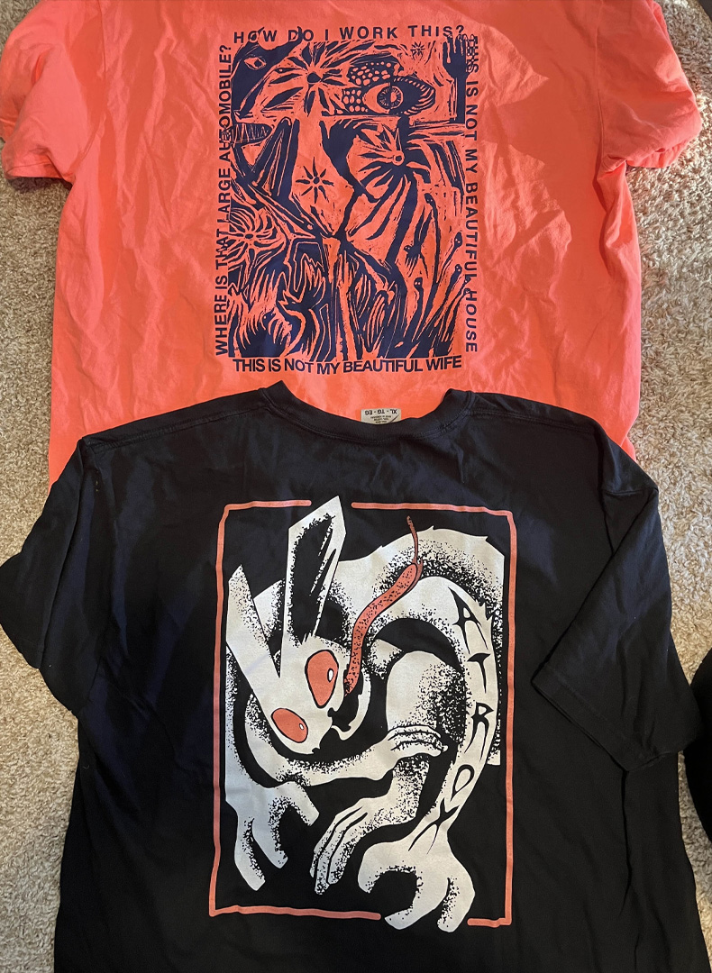 Two t-shirts, the top one is pink with a rough illustration and the text How do I work This? This is not my beautiful house. This is not my beautiful wife. Where is that large automobile? and the bottom is a rabbit illustration with long limbs and its tongue sticking out.